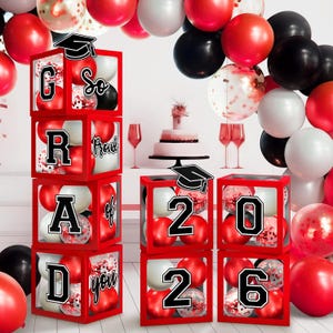 Könnte beinhalten: Rote Boxen mit roten, weißen und schwarzen Luftballons bilden die Wörter "GRAD" und "2026" für eine Abschlussfeier. Die Boxen sind mit Abschlusskappen verziert und von Luftballons und einer Torte umgeben.