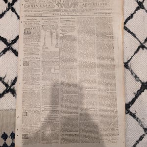 以下が含まれることがあります： 1797年3月発行の古新聞、Independent Chronicle and Universal Advertiser。古くなった紙は、小さなテキストと装飾的な見出しが並んだ、色あせた外観をしています。新聞は模様のある表面に置かれています。