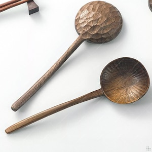 Puede incluir: Dos cucharas de madera con mangos largos. Una cuchara tiene un cuenco texturizado y tallado, mientras que la otra tiene un cuenco liso y acanalado. Las cucharas son de color marrón cálido y parecen hechas a mano.