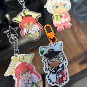 May include: Collection of anime-style acrylic keychains featuring colorful characters. Keychains include a character with red hair and a pumpkin, a character with white hair and a pink outfit, and a character with a white hat.
