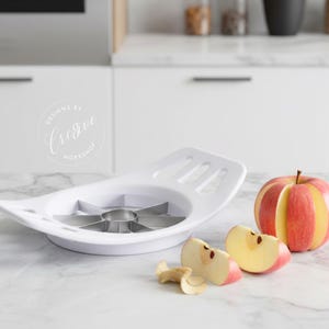 Puede incluir: Cortador de manzanas blanco con cuchillas de acero inoxidable junto a una manzana roja y amarilla y rodajas de manzana. El cortador tiene el texto "Designed by Cregee Workshop".