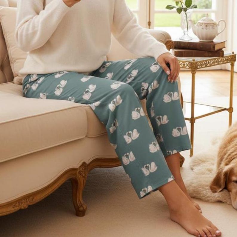 Puede incluir: Pantalones de pijama color turquesa con un estampado de cisnes blancos. Los pantalones est&aacute;n hechos de un material suave y los lleva una persona sentada en un sof&aacute; beige. La persona lleva un su&eacute;ter blanco.