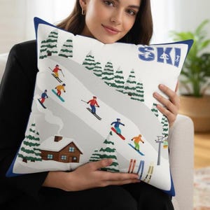 May include: White square pillow with a blue border featuring a ski resort scene. The design includes skiers, snow-covered slopes, evergreen trees, and a cabin. The word "SKI" is printed in blue.