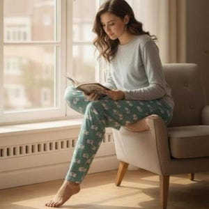 Puede incluir: Una persona con una camisa de manga larga gris claro y un pantal&oacute;n de pijama estampado, sentada en un sill&oacute;n beige y leyendo un libro. El pantal&oacute;n de pijama tiene un fondo verde azulado claro con un estampado floral blanco. La habitaci&oacute;n tiene una ventana y un radiador.