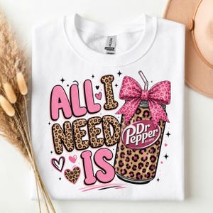 May include: A white t-shirt featuring a graphic print with the text "ALL I NEED IS" in pink and leopard print lettering. Next to the text is a soda can with a leopard print pattern, topped with a large pink leopard print bow and a straw.