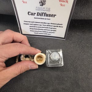 May include: A clear glass car diffuser bottle with a light-colored wooden cap and a hanging cord, accompanied by a white instruction card labeled Woodland Aromas Black Ice Car Diffuser with text detailing usage instructions and safety warnings.