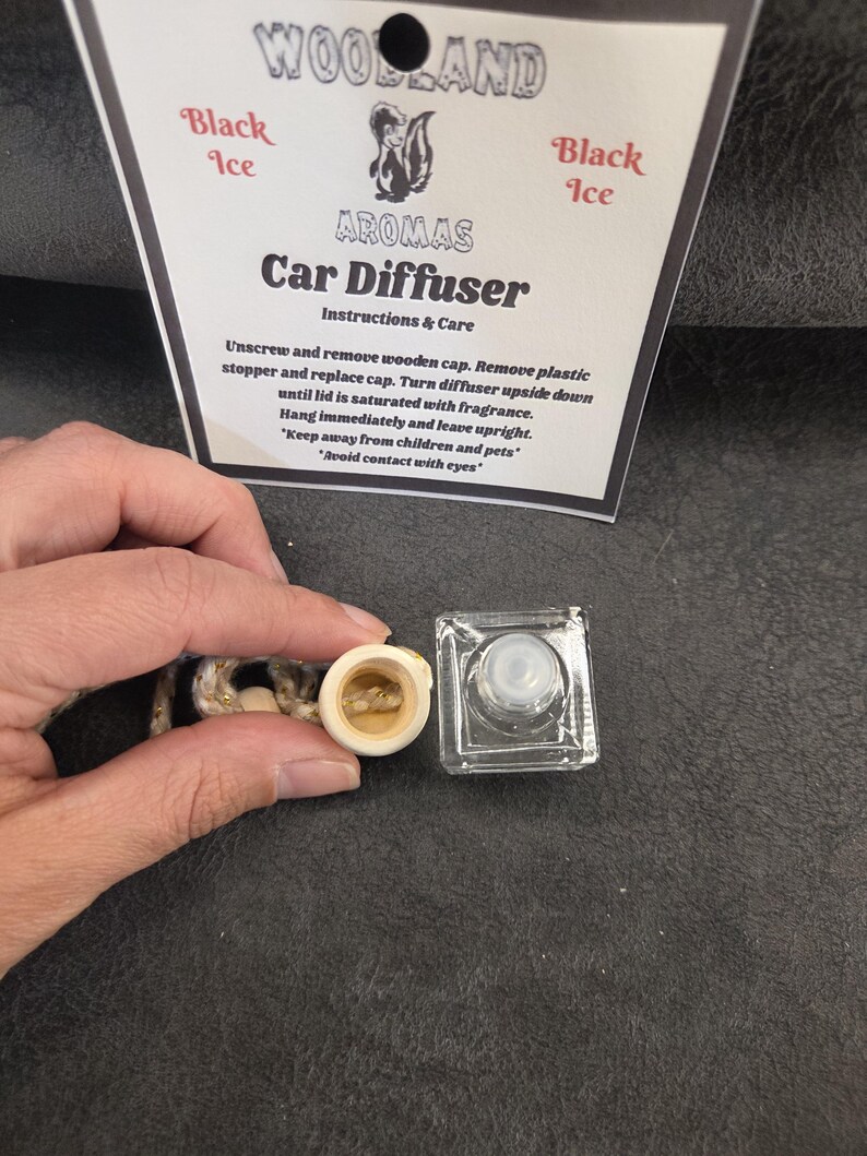 May include: A clear glass car diffuser bottle with a light-colored wooden cap and a hanging cord, accompanied by a white instruction card labeled Woodland Aromas Black Ice Car Diffuser with text detailing usage instructions and safety warnings.