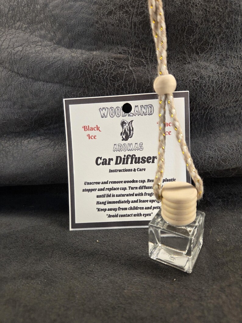 May include: A clear glass car diffuser bottle with a light-colored wooden cap and a beige hanging cord. Behind the bottle is a white instruction card labeled Woodland Aromas Car Diffuser with a skunk graphic and Black Ice scent text.