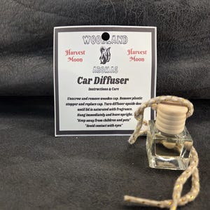 May include: A clear glass car diffuser with a light-coloured wooden cap and a beige cord with gold flecks. Behind it is a white product tag labelled Woodland Aromas Harvest Moon Car Diffuser with printed instructions and care information.