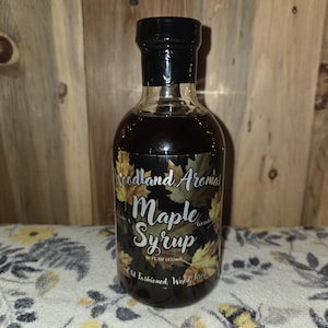 May include: A clear glass bottle of maple syrup with a black cap. The label reads "Woodland Aromas Maple Syrup" with leaf illustrations. The bottle contains 16 fl oz (473ml) of syrup.
