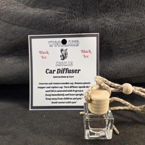 May include: A clear glass car diffuser bottle with a light wooden cap and a braided beige cord. Beside it is a white instruction card labeled Woodland Aromas Car Diffuser Black Ice, featuring a skunk graphic and text detailing usage instructions.