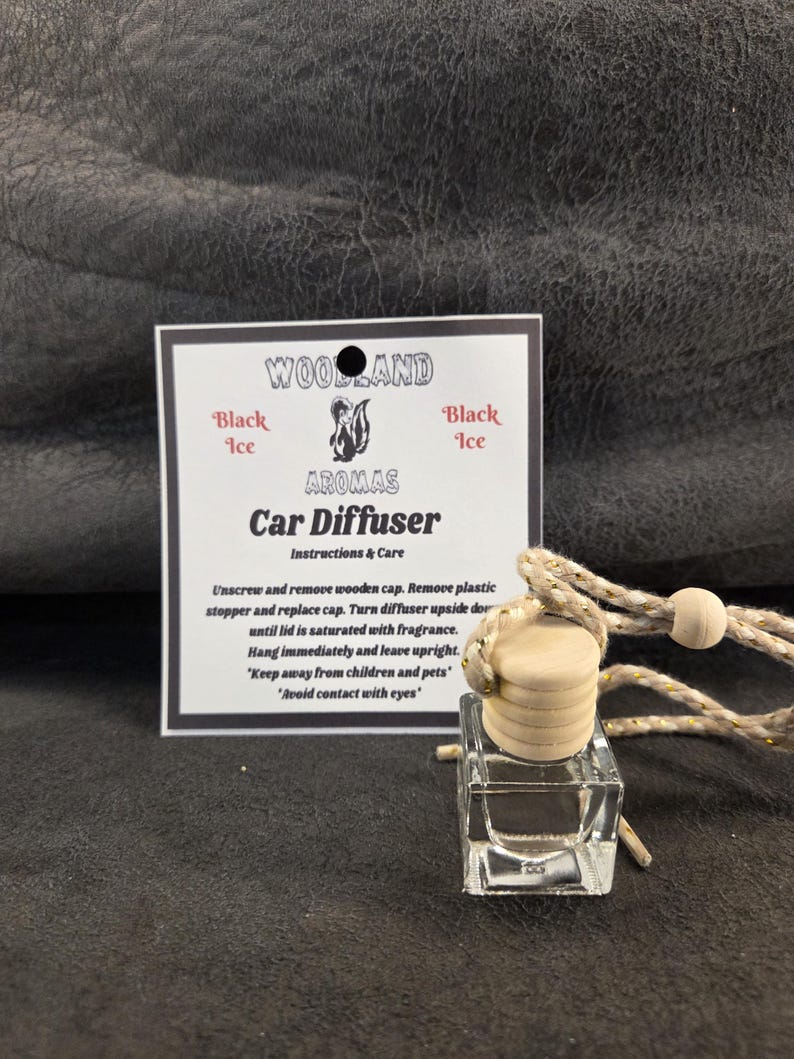 May include: A clear glass car diffuser bottle with a light wooden cap and a braided beige cord. Beside it is a white instruction card labeled Woodland Aromas Car Diffuser Black Ice, featuring a skunk graphic and text detailing usage instructions.