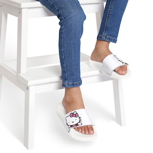 Op de afbeelding: Witte slippers met een afbeelding van het Hello Kitty-personage met een roze strik op de band. De slippers hebben een brede, platte witte zool en een enkele brede band over de bovenkant van de voet, gedragen met een blauwe spijkerbroek.