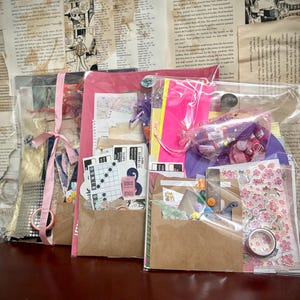 Peut inclure: Quatre kits de bricolage dans des sacs en plastique transparents et des enveloppes en papier brun. Chaque kit contient diverses fournitures de bricolage, notamment des autocollants, des boutons, du ruban washi et du papier décoratif. Les kits sont attachés avec des rubans roses.