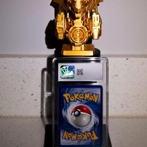 May include: A gold-colored, stylized dragon figure sits atop a graded Pokémon card in a protective case. The card features the Pokémon logo and a Poké Ball design on a blue background. The dragon figure has intricate details.