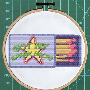 Może przedstawiać: Haft krzyżykowy w drewnianym tamborku. Projekt przedstawia pudełko zapałek z napisem "GOT MY SPARK BACK" i żółtą gwiazdą. Zapałki są różowe i żółte. Tło to zielona kratka.