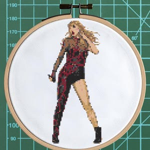 May include: A cross-stitch embroidery of a person in a performance outfit, set within a wooden hoop. The figure is depicted in a red and black patterned bodysuit, with blonde hair and black boots. The background is white, and the hoop is set against a green grid.