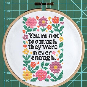 Puede incluir: Bordado de punto de cruz en un aro de madera. El diseño presenta el texto "You're not too much they were never enough" en negro, rodeado de flores coloridas en rosa, naranja, amarillo y morado. El fondo es blanco.
