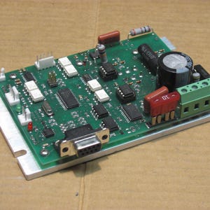 Puede incluir: Una placa de circuito verde con varios componentes electrónicos, incluyendo chips, resistencias y un gran condensador. La placa está montada sobre una base de metal plateado y presenta un bloque de terminales verde y un conector negro.
