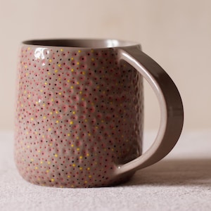 Könnte beinhalten: Eine braune Keramiktasse mit einer strukturierten Oberfläche und einem gesprenkelten Muster in Rot-, Gelb- und Schwarztönen.