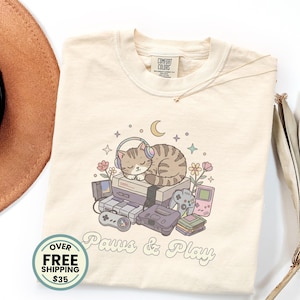 May include: A cream-colored t-shirt featuring a graphic of a sleeping tabby cat wearing headphones, resting on a stack of vintage video game consoles. The design includes flowers, stars, a crescent moon, and the text Paws & Play in a cursive font.