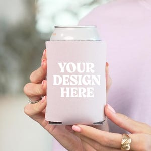 Puede incluir: Un enfriador de latas rosa claro con el texto blanco "YOUR DESIGN HERE". El enfriador sostiene una lata plateada. La imagen es de un portavasos.
