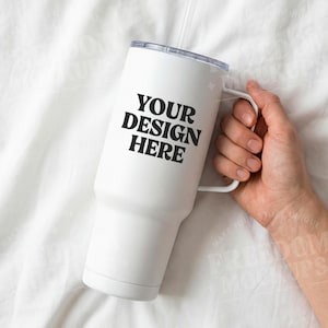 May include: A white tumbler with a handle and a clear straw. The text "YOUR DESIGN HERE" is printed in black on the front of the tumbler. The tumbler is held by a person's hand.
