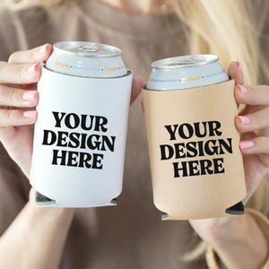 Puede incluir: Dos fundas para latas de bebidas, una blanca y otra beige, cada una con el texto "YOUR DESIGN HERE" en letras negras en negrita. Las fundas están diseñadas para latas de tamaño estándar. La imagen muestra las fundas sostenidas por una persona.