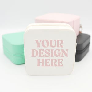 May include: A collection of square, zippered jewelry cases in various pastel colors. The front-most case is white with the text "YOUR DESIGN HERE" in pink. Other cases are mint green, pink, and black.