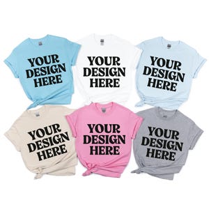 May include: Six short-sleeved t-shirts in various colors: light blue, white, light blue, beige, pink, and gray. Each shirt features the text "YOUR DESIGN HERE" in bold black letters, perfect for custom designs.