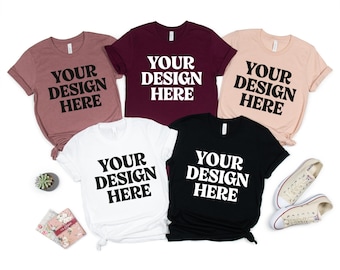 May include: Five t-shirts in various colors: mauve, burgundy, peach, white, and black. Each shirt has the text "YOUR DESIGN HERE" printed in a bold, white font. A pair of white canvas sneakers and small decorative items are also present.