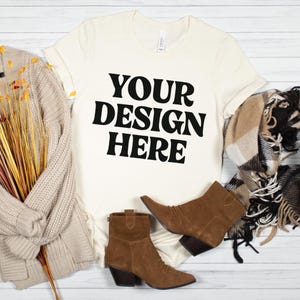 May include: Cream-colored t-shirt with the text "YOUR DESIGN HERE" in bold black letters. The shirt is styled with brown suede ankle boots, a beige sweater, and a patterned scarf. The items are arranged on a white wooden surface.