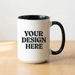 May include: White ceramic mug with a black interior and handle. The mug features the text "YOUR DESIGN HERE" in bold black letters. The mug is sitting on a wooden surface.