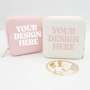 Może przedstawiać: Dwa kwadratowe etui na biżuterię, jedno różowe i jedno kremowe, z napisem "YOUR DESIGN HERE" wydrukowanym na przodzie. Złota bransoletka z zawieszkami znajduje się przed etui. Etui mają zamki błyskawiczne.