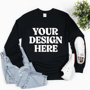 May include: Black long-sleeve shirt with the text "YOUR DESIGN HERE" in white. The shirt is paired with light blue jeans and white sneakers. A small green plant is in the background.