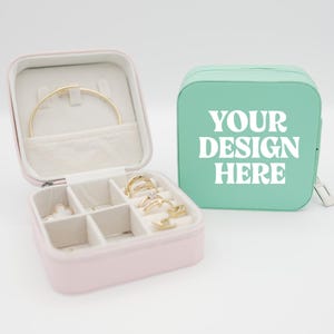 Puede incluir: Un estuche de viaje para joyas rosa, abierto para mostrar una pulsera y anillos dorados. Un estuche verde menta separado con el texto "YOUR DESIGN HERE" está al lado. El estuche de joyería tiene múltiples compartimentos.
