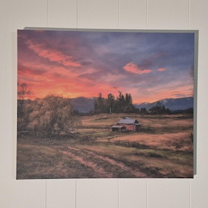 Könnte beinhalten: Gerahmtes Landschaftsgemälde, das einen Sonnenuntergang über einer ländlichen Szene zeigt. Der Himmel ist eine lebendige Mischung aus Rot, Orange und Lila. Eine kleine rote Scheune steht auf einem Feld, mit Bäumen und Bergen im Hintergrund. Das Kunstwerk wird an einer weißen Wand ausgestellt.