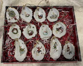 May include: A set of twelve oyster shell Christmas ornaments. Each white shell is hand-painted with a different holiday scene and a gold-painted edge. The ornaments are displayed in a red and white box with red and white paper filler.