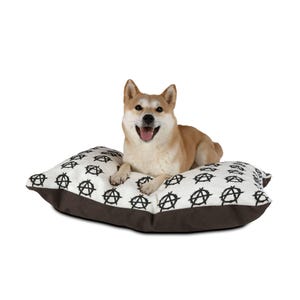 May include: A Shiba Inu dog rests on a dog bed. The bed is rectangular, with a white top featuring a repeating black anarchy symbol pattern. The bed's bottom and sides are brown.