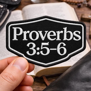 May include: A black patch with white embroidered text reading "Proverbs 3:5-6". The patch has a white border and a shield shape. The patch is held in a hand, with a leather jacket, open book, candle, and cross in the background.