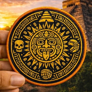 May include: Round embroidered patch with a black background and a yellow Aztec sun design. The patch has an orange border and features a skull, a stylized face, and other geometric shapes. The background shows a pyramid and a sunset.