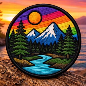 May include: Circular embroidered patch featuring a mountain landscape with a river, trees, and a sunset. The mountains are blue with white snow caps. The sky is a rainbow of colors. The patch is on a wooden log.
