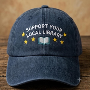 May include: A navy blue baseball cap with a distressed finish. The front features white embroidered text that reads SUPPORT YOUR LOCAL LIBRARY, accented by six small yellow embroidered stars and a small open book icon in white and teal.
