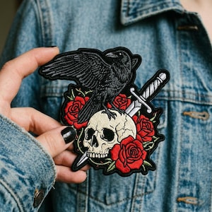 May include: Embroidered patch featuring a black raven perched atop a skull, surrounded by red roses. A silver dagger is positioned behind the skull. The patch is black, red, white, and silver.