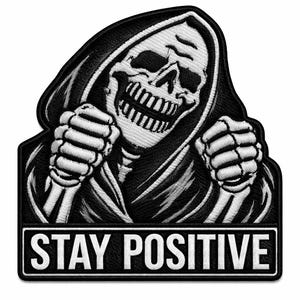 May include: Embroidered patch featuring a grinning skull in a black hooded robe, with skeletal hands clenched in fists. The phrase "STAY POSITIVE" is displayed in white block letters on a black rectangular banner below the skull.