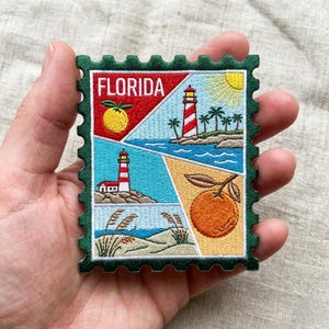 May include: Embroidered patch in the shape of a postage stamp featuring Florida imagery. The design includes a red and white striped lighthouse, an orange, a sun, and the word "FLORIDA". The patch has a green border.