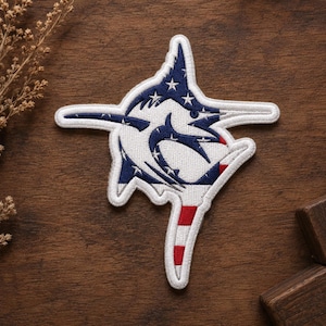May include: Embroidered patch featuring a marlin design with an American flag motif. The patch has a white outline, with the marlin's body filled with a white background, blue stars, and stripes. The tail has red and white stripes.