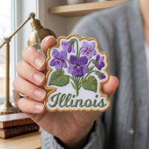 May include: An embroidered patch featuring a cluster of purple violet flowers with green leaves on a white background. The word Illinois is stitched in green cursive script at the bottom. The patch has a scalloped gold border edge.