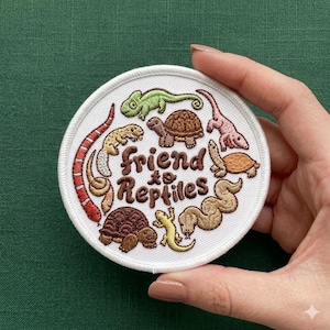 May include: A circular white embroidered patch featuring various reptiles including a green chameleon, brown turtles, a red and white snake, and a yellow lizard. The text "friend to Reptiles" is stitched in brown thread in the center of the patch.