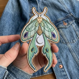 May include: Embroidered moth patch featuring a crescent moon design. The patch has a light green and gold color scheme with accents of purple and blue. The patch is held against a denim jacket.